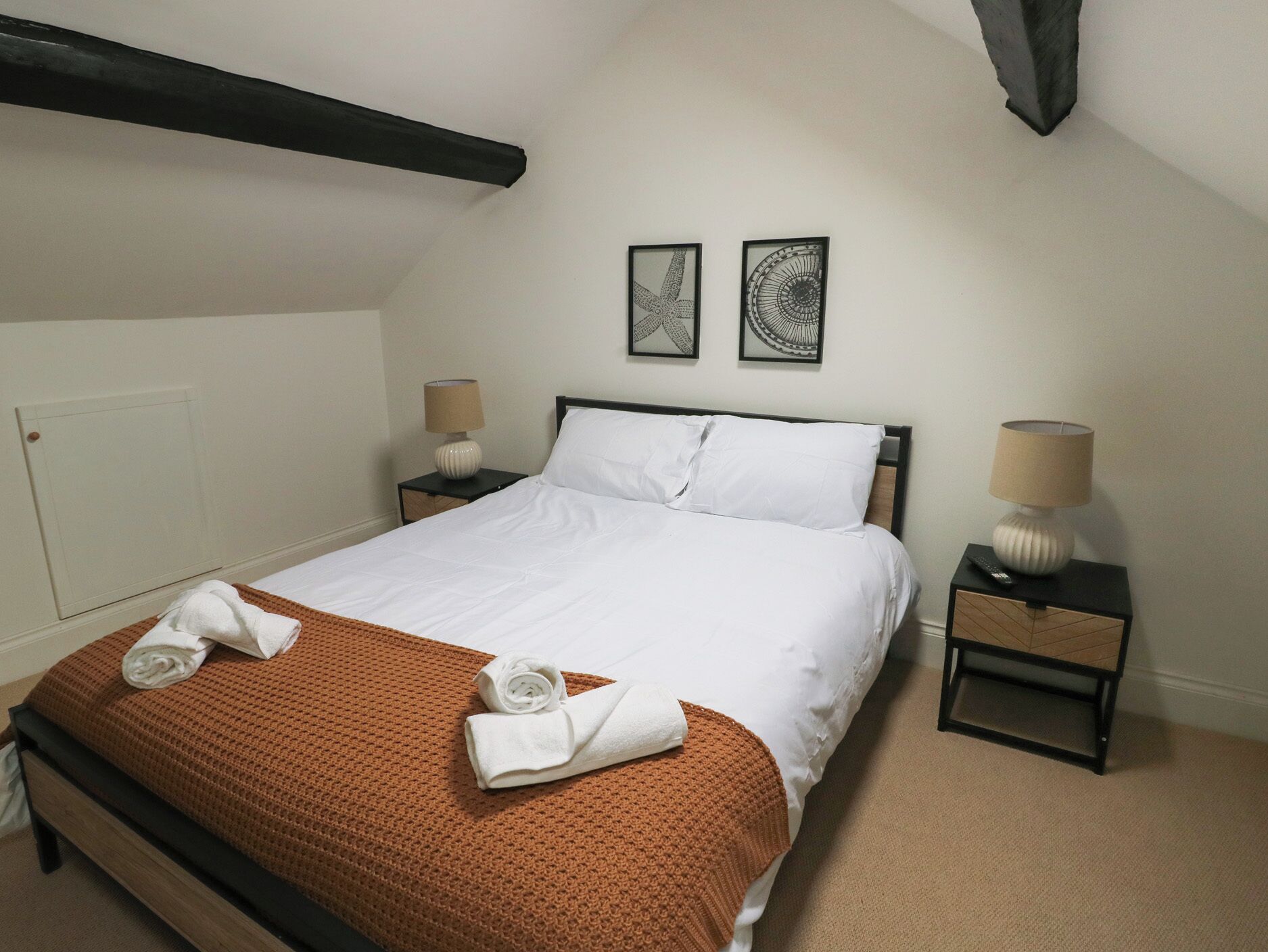 A neatly made bed with white linens, a brown knitted blanket, and two rolled towels on the foot of the bed. There are two bedside tables with lamps and framed artwork on the wall above the bed.
