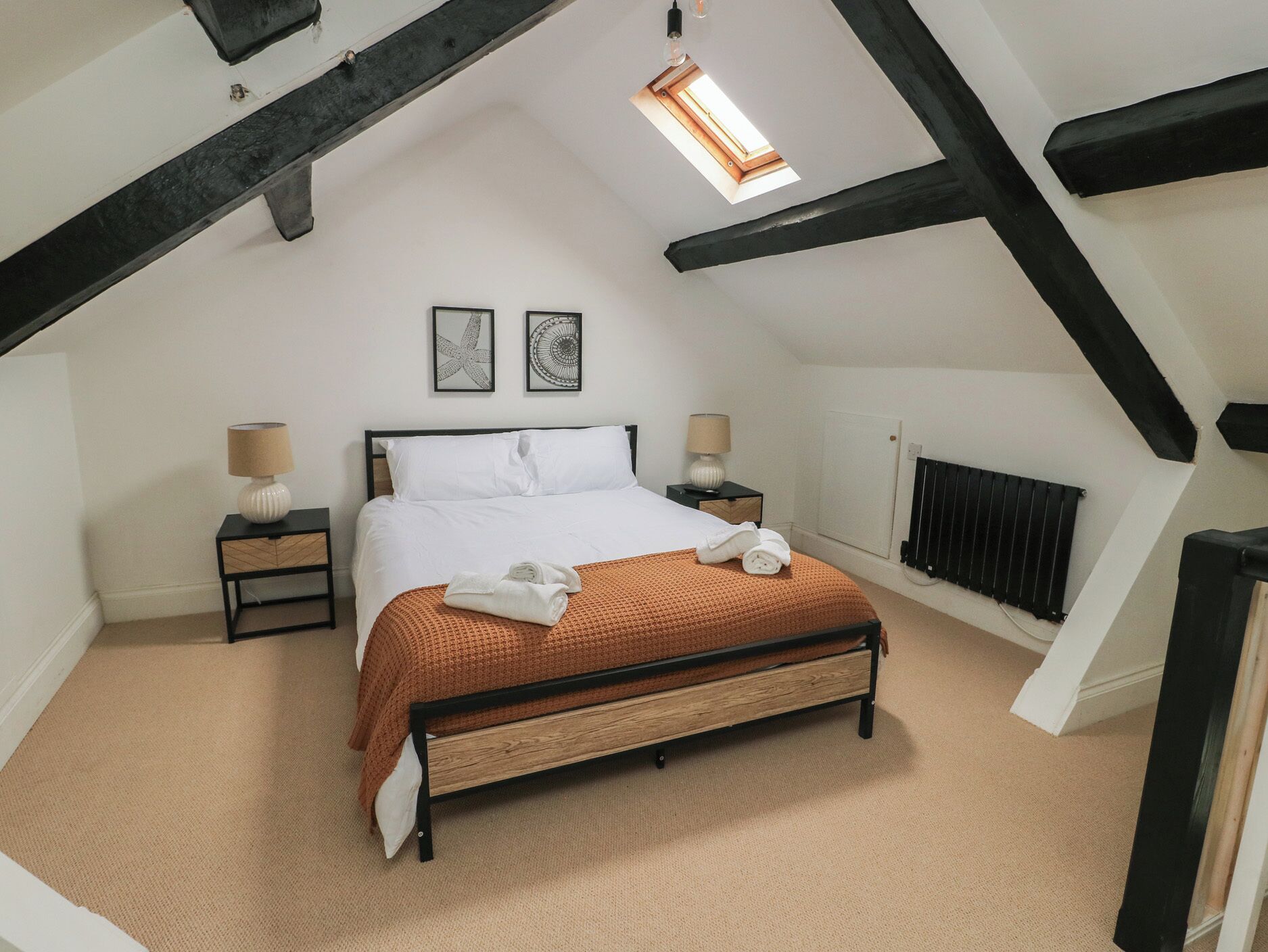 A bedroom with a sloped ceiling, a bed with a brown blanket, two bedside lamps, and a radiator.