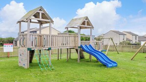 A playground with a slide and climbing net, surrounded by a grassy area and wooden play structures. - Skylarks! (St. Agnes)