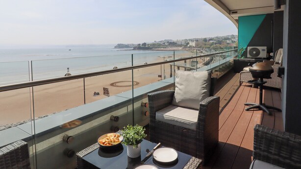 Un balcón con vistas a la playa, muebles de mimbre, una mesa con platos y un bol de comida, y un equipo de aire acondicionado.