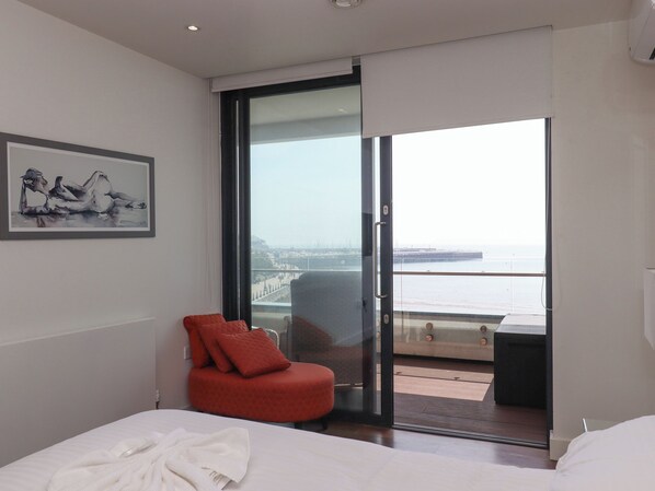 A modern hotel room with a bed, a red chair, a glass door leading to a balcony with a view of the sea and a bridge.