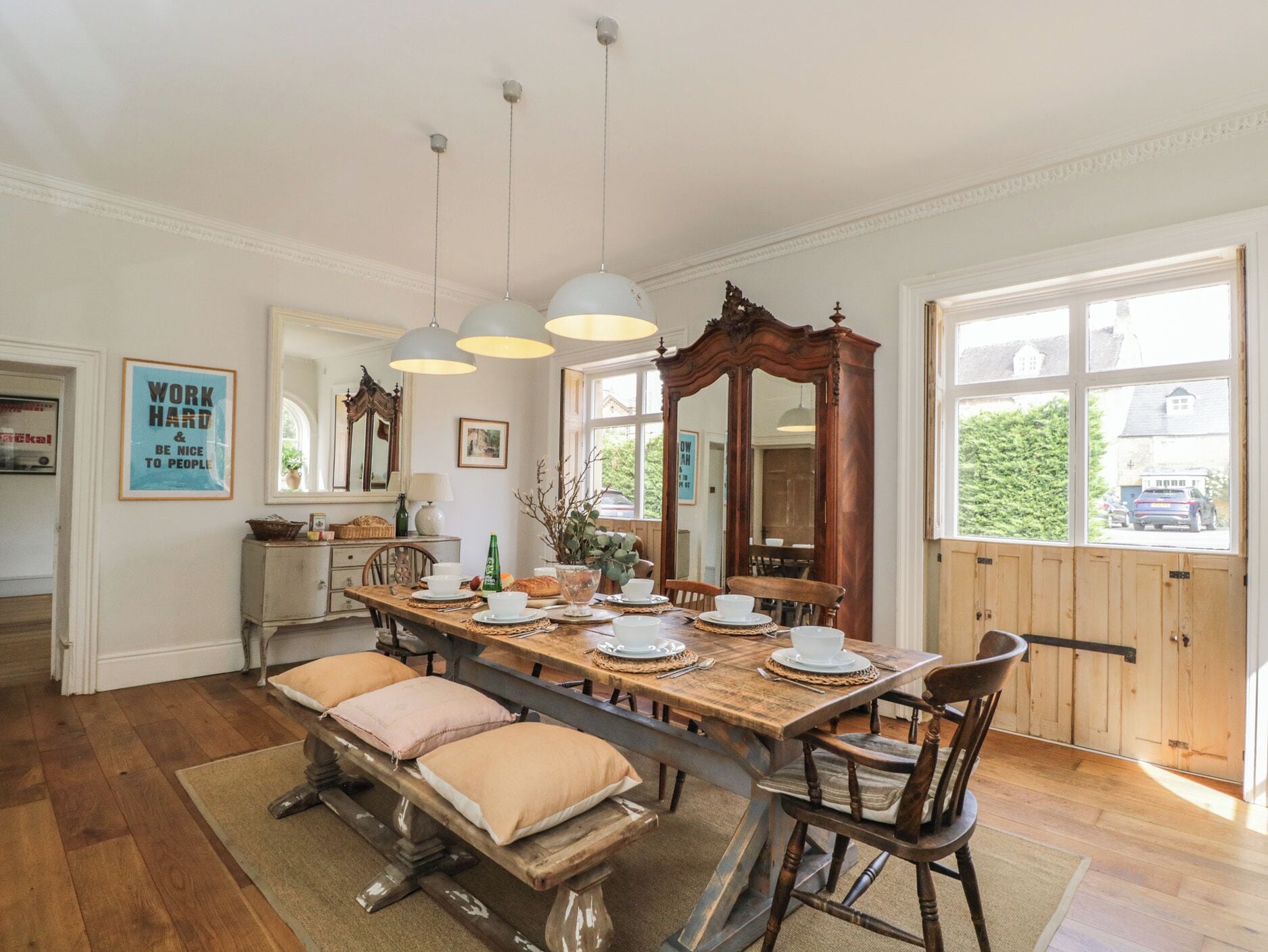 Uma sala de jantar com mesa de madeira posta para quatro pessoas, um espelho grande e luminárias pendentes.