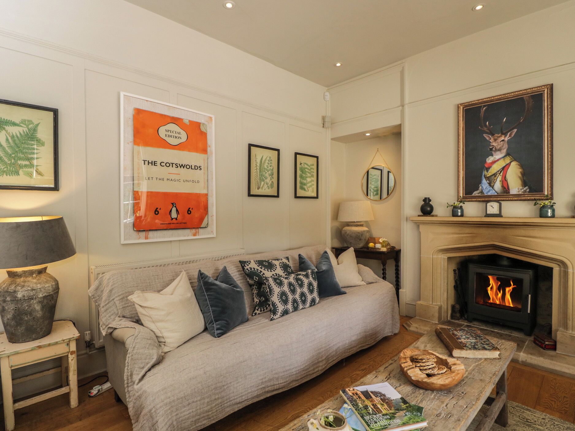A cozy living room with a fireplace, a sofa with patterned cushions, a coffee table with books, and framed botanical prints on the wall.