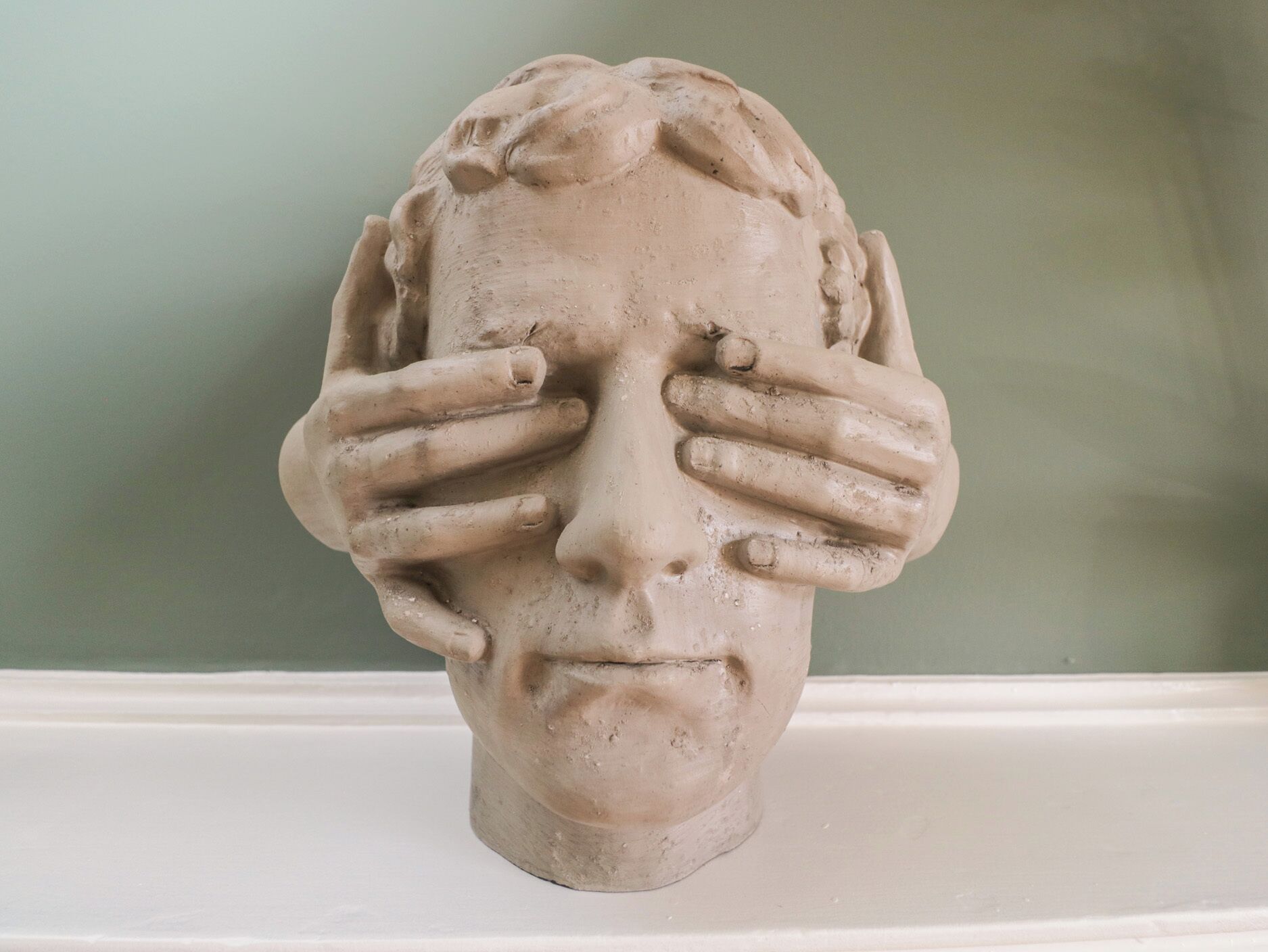 A sculpture of a human head with hands covering the face, set against a plain background.