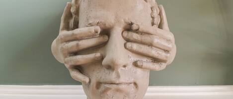 A sculpture of a human head with hands covering the face, set against a plain background.