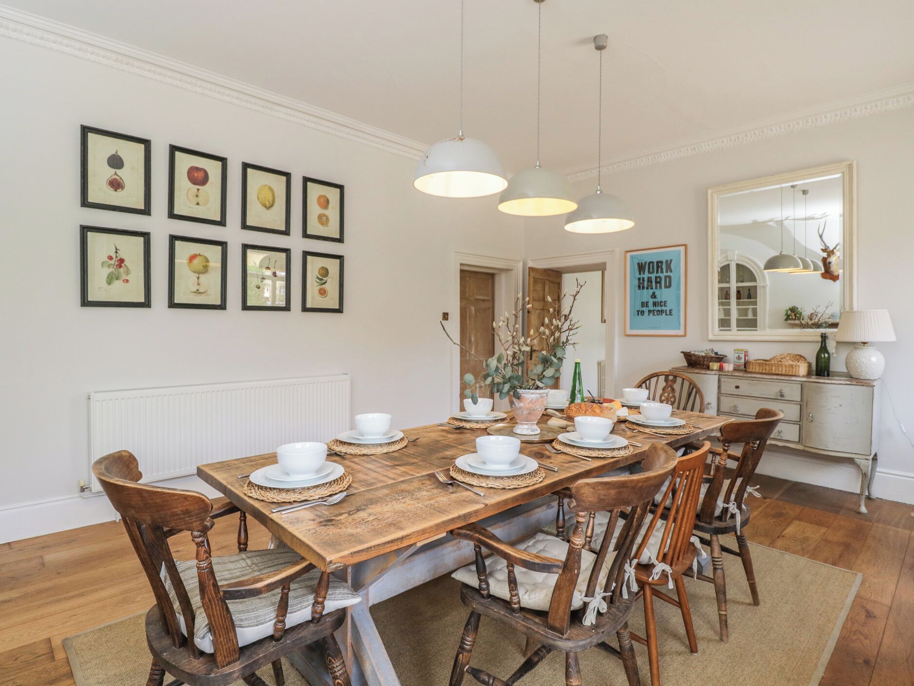 Une salle à manger avec une table en bois dressée pour six personnes, entourée de six chaises. Au mur, au-dessus de la table, on peut voir des reproductions encadrées de gravures botaniques.