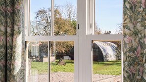 A room with a large window divided into four panes, offering views of a garden and a building with a curved roof. - Elm Tree Cottage (Thirsk)