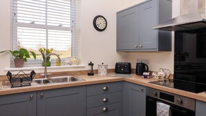 A modern kitchen with grey cabinets, a stainless steel sink, a black stove, and a window with blinds. - Elm Tree Cottage (Thirsk)