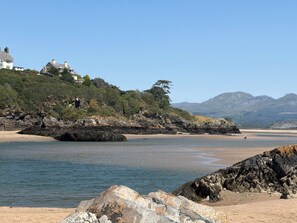 Paisagem costeira com uma praia de areia, afloramentos rochosos e uma colina com casas.