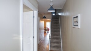 Un couloir avec un plancher en bois, une porte blanche, un escalier et une suspension lumineuse.