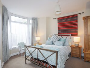 A bedroom with a bed, two bedside tables, a chair, a window with curtains, and a red patterned wall hanging. - The Quarterdeck (Porthmadog)