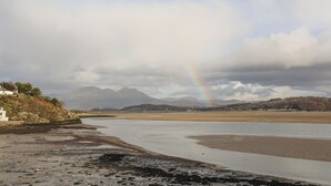 Une rivière où l’on aperçoit un arc-en-ciel, des montagnes en arrière-plan et un bâtiment sur la gauche.