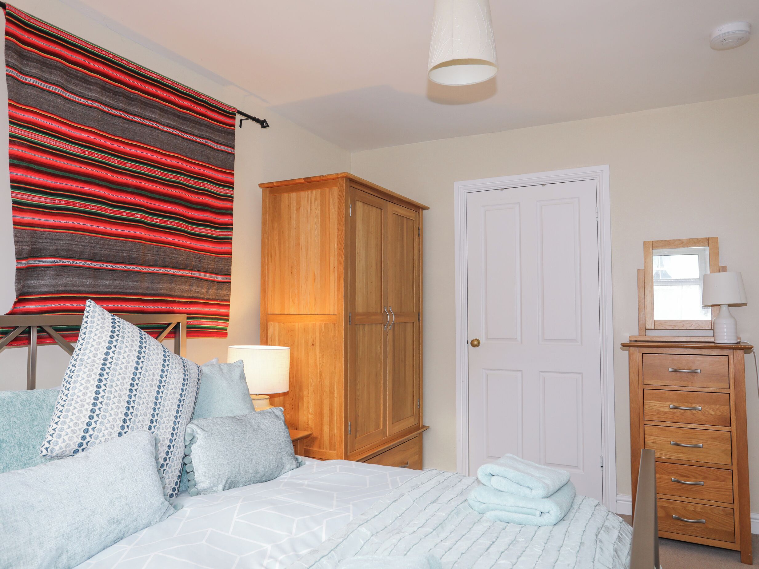 A bedroom with a bed, a wooden wardrobe, a white door, a bedside table with a lamp, and a red and black striped blanket.