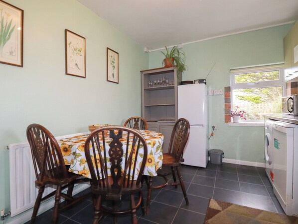 A dining area with a table covered by a floral tablecloth, surrounded by wooden chairs. A white refrigerator and a washing machine are visible in the background. - Bramble Cottage (Hayle)