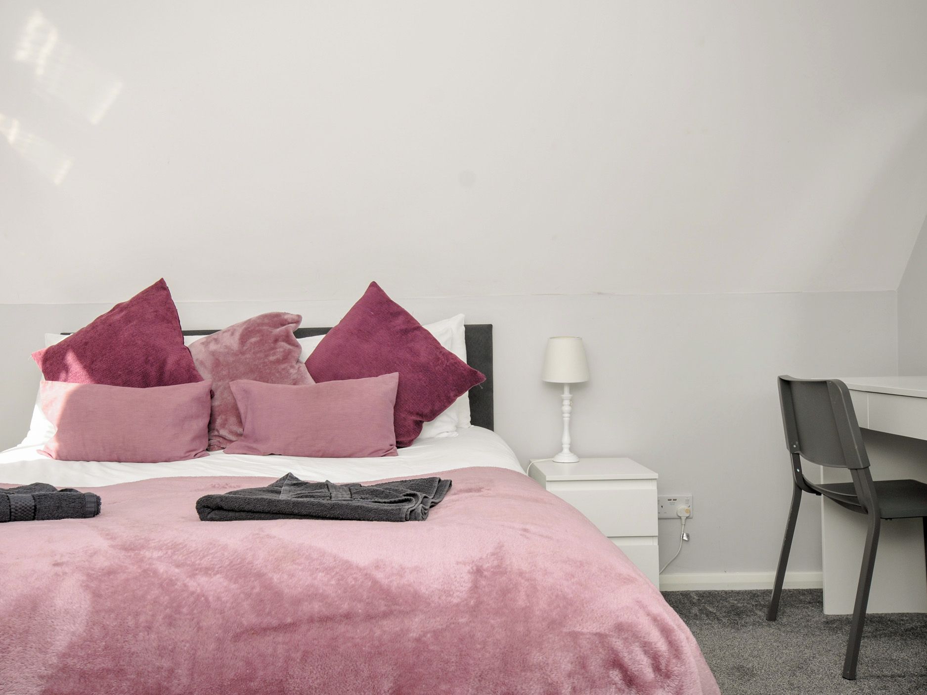 Un dormitorio con una cama, almohadas, una toalla doblada, una mesita de noche con una lámpara y una silla.