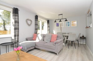 A modern living room with a grey sofa, a dining table with chairs, and a window with curtains. - Lewis Lodge (Gunnislake)
