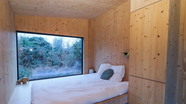 A wooden cabin bedroom with a large window, a bed with pillows, and a wicker chair. - Broc (Kinvara)