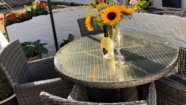 Une table ronde en verre, des chaises en osier, un vase rempli de fleurs et une bouteille de vin.