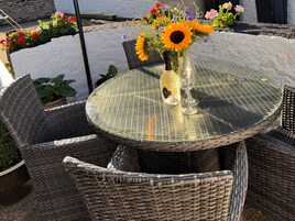 Uma mesa de vidro redonda com cadeiras de vime, um vaso com flores e uma garrafa de vinho.