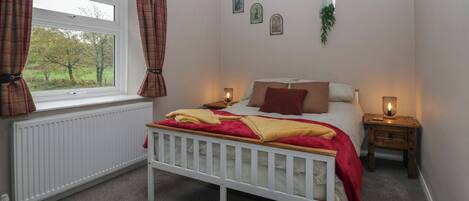 Ein Schlafzimmer mit weißem Babybett, einem Fenster mit Karottenvorhängen, einem Nachttisch mit Lampe und einem Heizkörper.