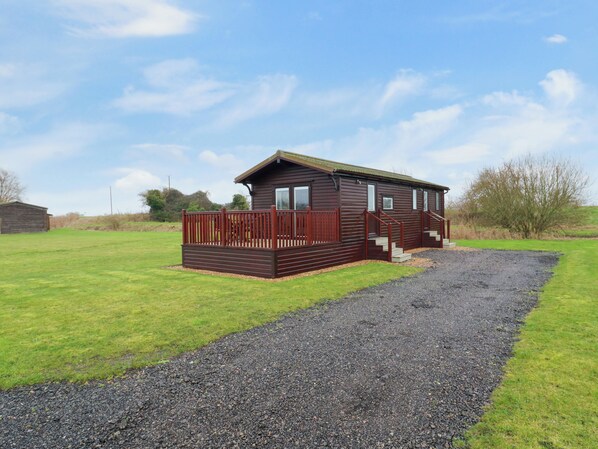 A small wooden cabin with a deck, surrounded by a gravel path and grassy area. - Cuckoo (Huntingdon)