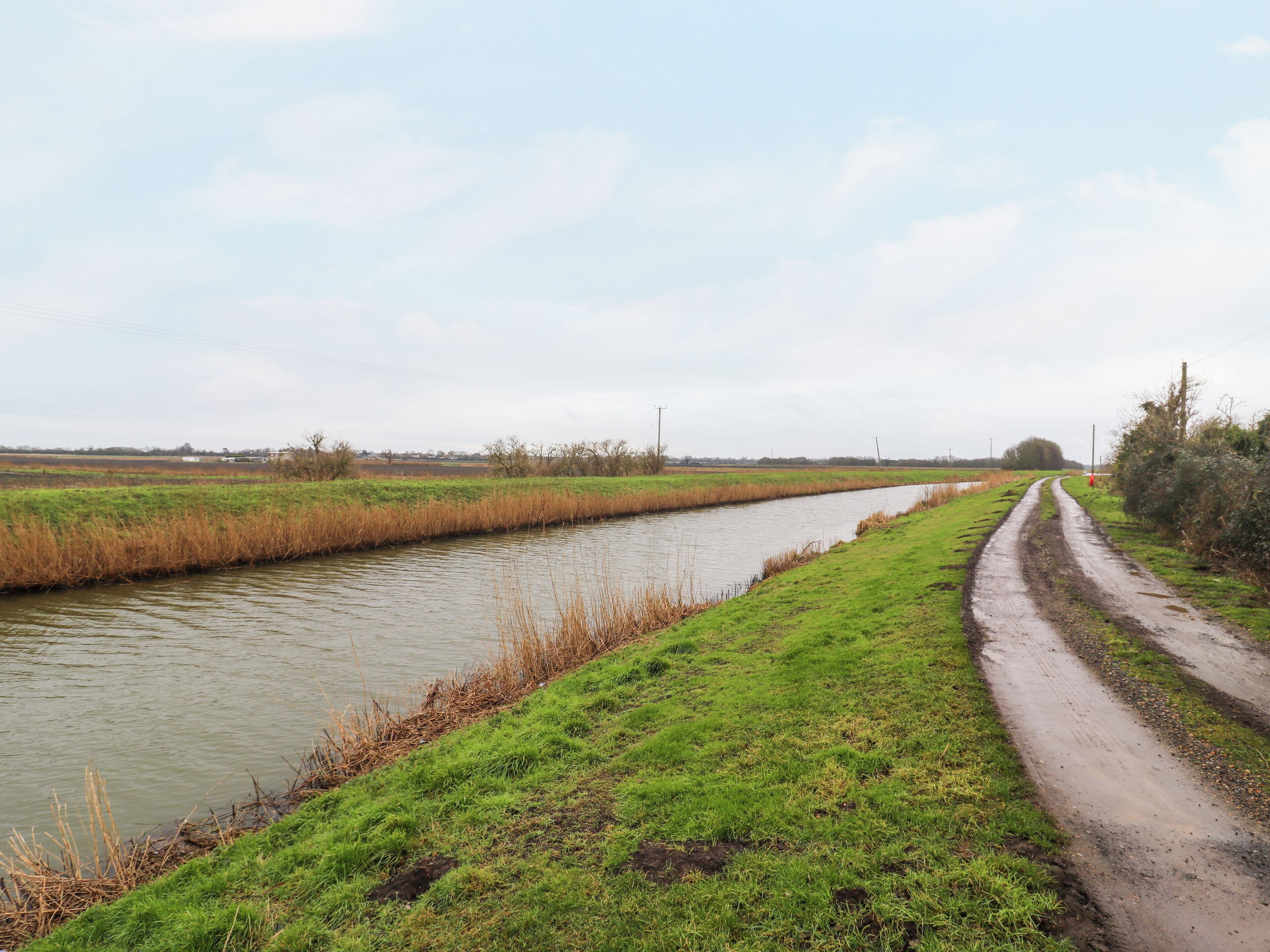 Un paysage rural avec un étroit canal d’eau, un sentier boueux et des champs ouverts.