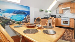 A dining area with a table set for four, wooden chairs, a view of a coastal landscape through a large window, and a kitchen with wooden cabinets and modern appliances. - 20 Keay Heights (St Austell)