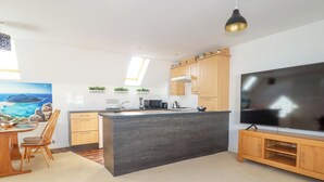 A modern kitchen with a central island, wooden cabinets, a large flat-screen TV, and a dining area with a wooden table and chairs. - 20 Keay Heights (St Austell)