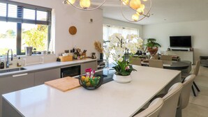 Una cocina moderna con una isla central, una zona de comedor con sillas, un televisor y un amplio ventanal con persianas.