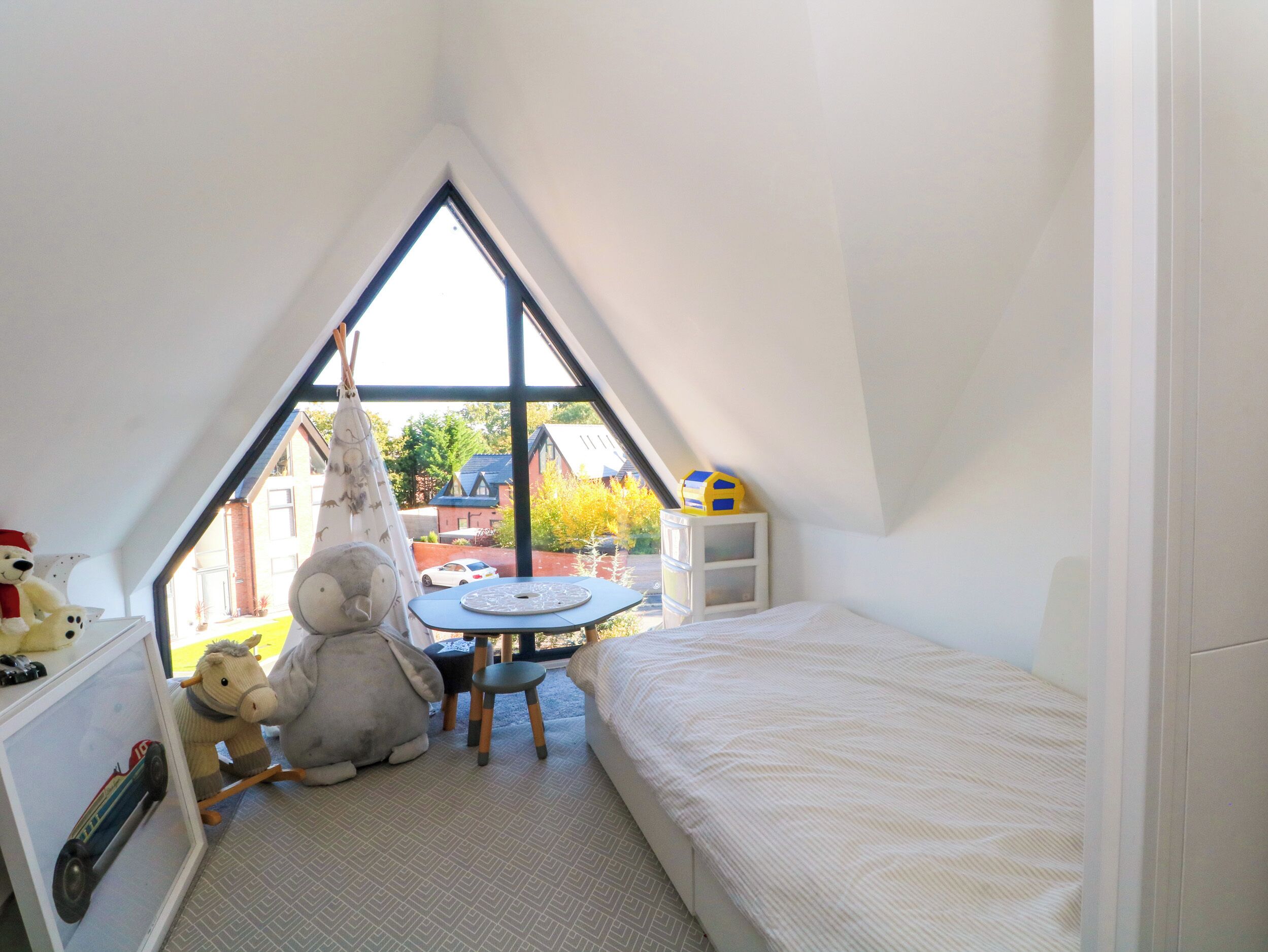 A child's bedroom with a sloped ceiling, a bed, a desk with toys, and a window overlooking a house.