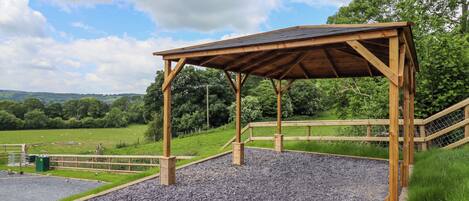 A wooden pavilion with a gravel floor, surrounded by a fence and trees.