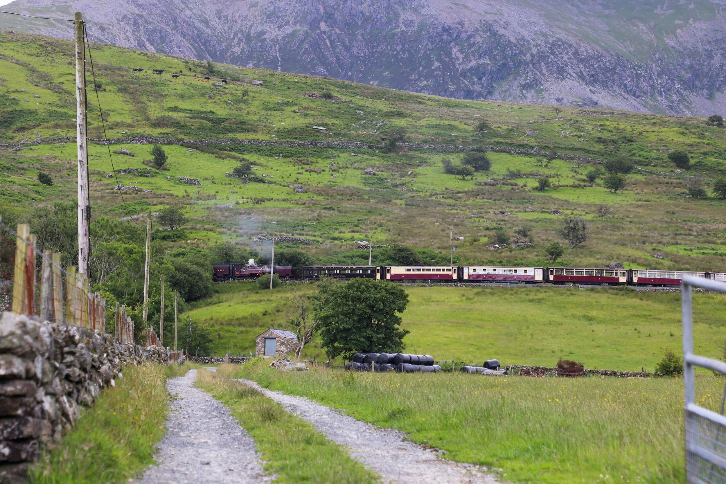 A train on a track through a rural landscape with green fields and a stone wall on the left.