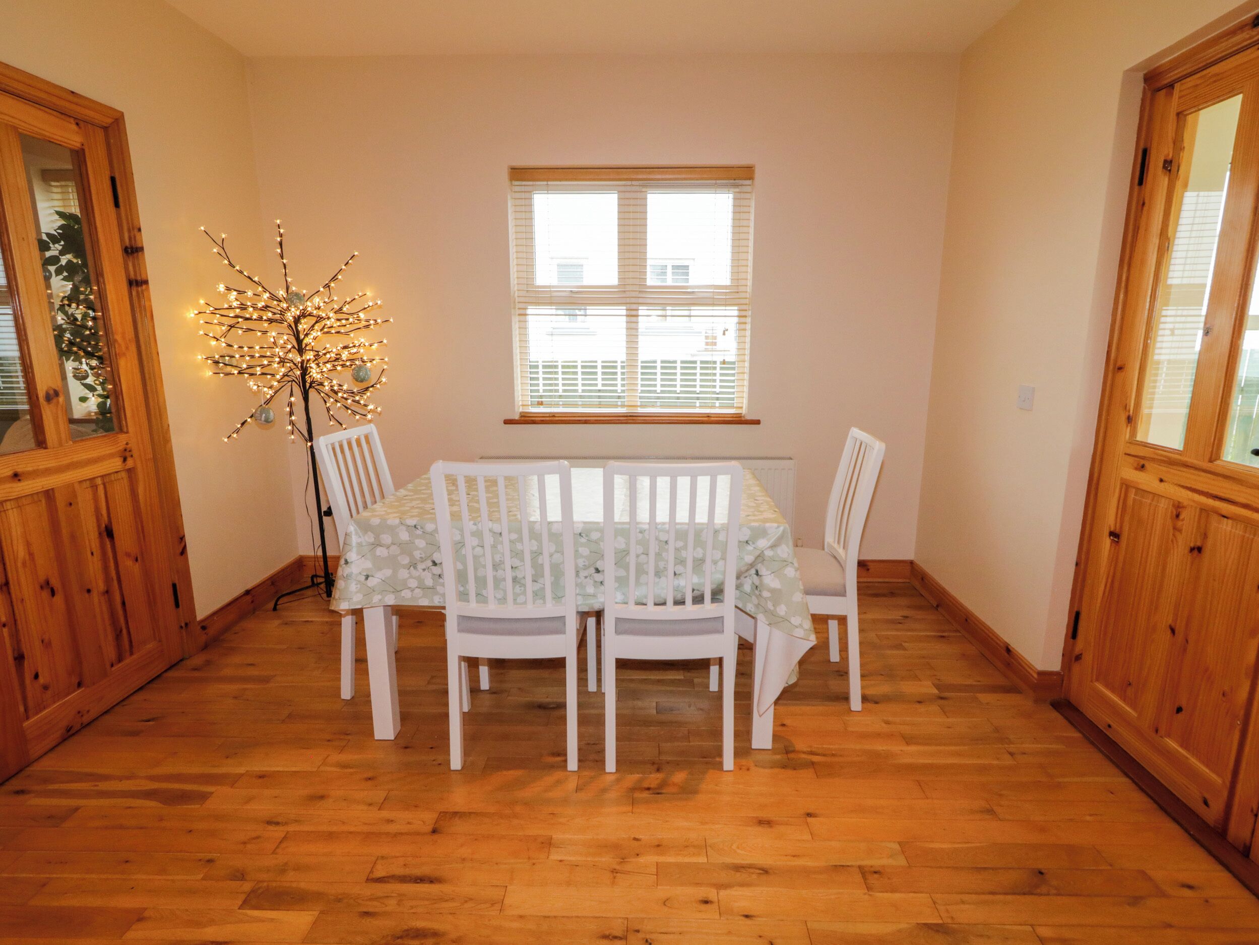 A dining room with a table set for four, white chairs, a wooden floor, and a window with blinds.