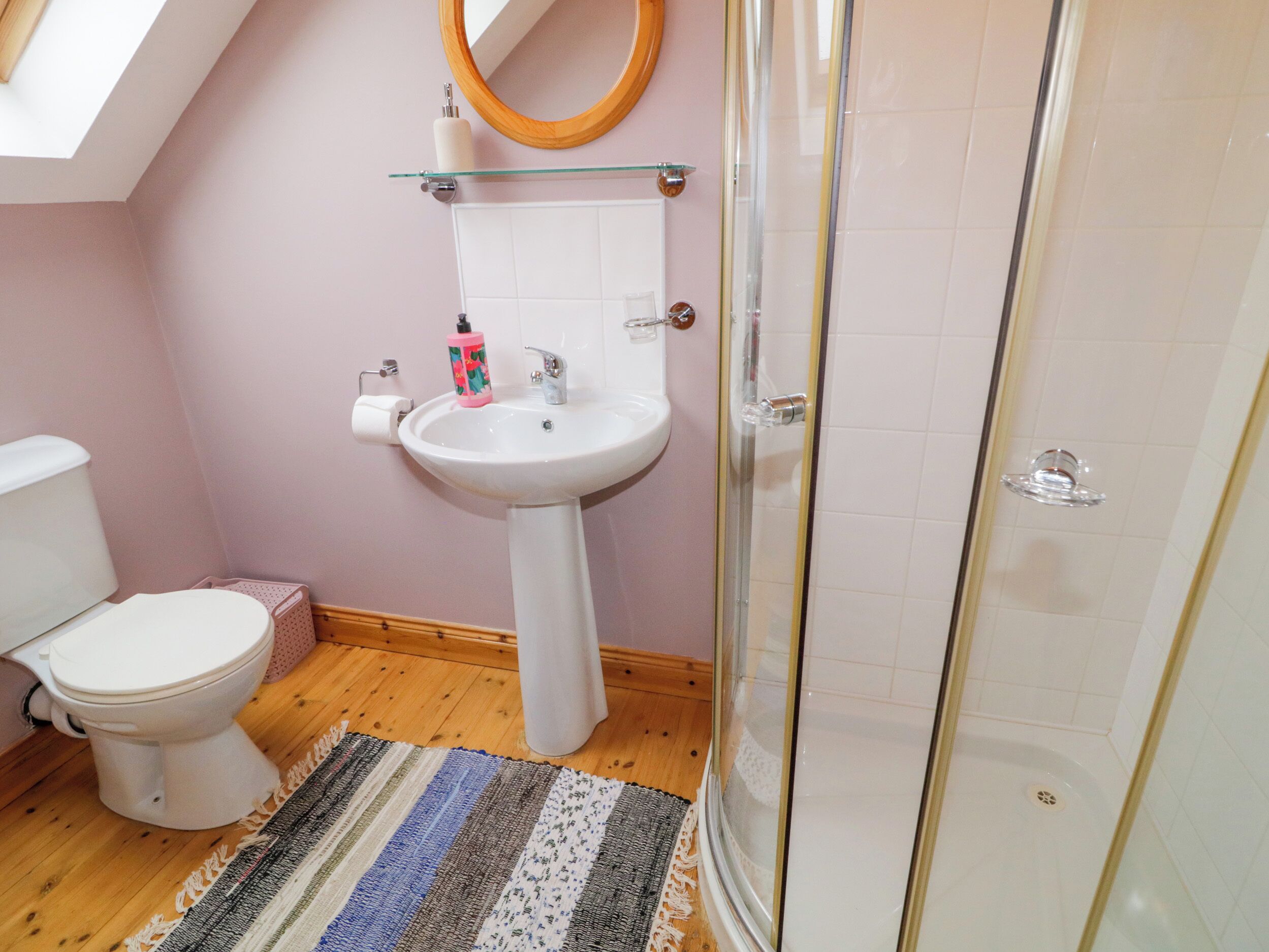 A bathroom with a white toilet, a pedestal sink, a glass shower enclosure, and a striped rug on the floor.