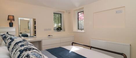 A bedroom with a bed, bedside table, lamp, mirror, windows, and a radiator.