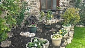 A stone building with a wooden window, surrounded by a garden with various potted plants and a stone pathway. - Rose Cottage (Scarborough)