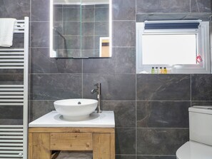 A bathroom with a white sink, wooden vanity, dark tiled walls, a mirror, and a window. - Calvin's Cottage (Arundel)