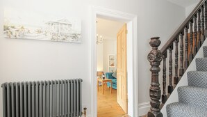 A hallway with a radiator, a wooden staircase, and a door leading to a room with a blue sofa and a lamp. - Albert House (Looe)