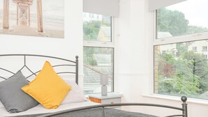 A bedroom with a bed, pillows, a bedside table, and a window with a view of buildings and trees. - Albert House (Looe)