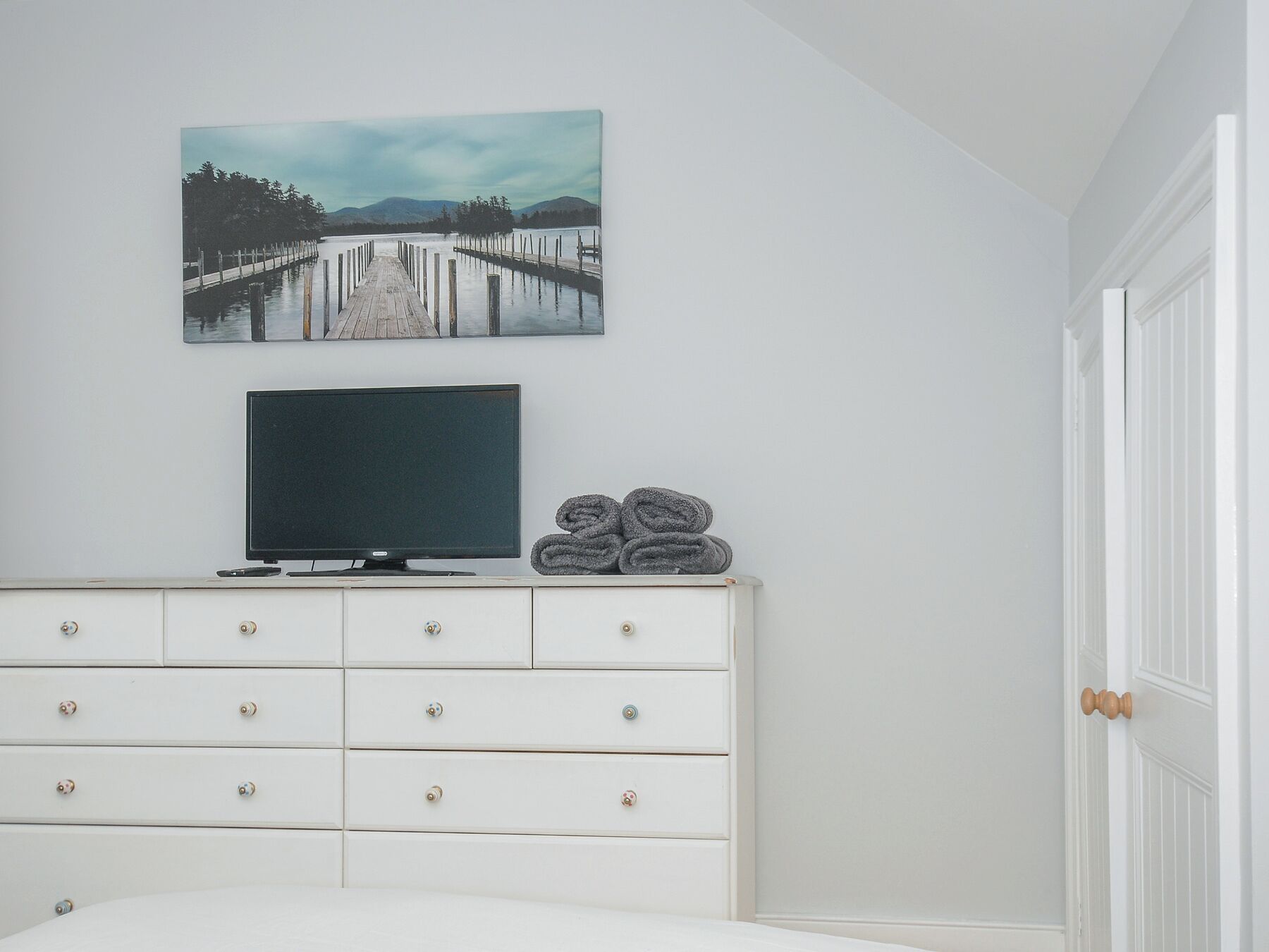 A bedroom with a white dresser, a flat-screen TV, and a painting of a pier.