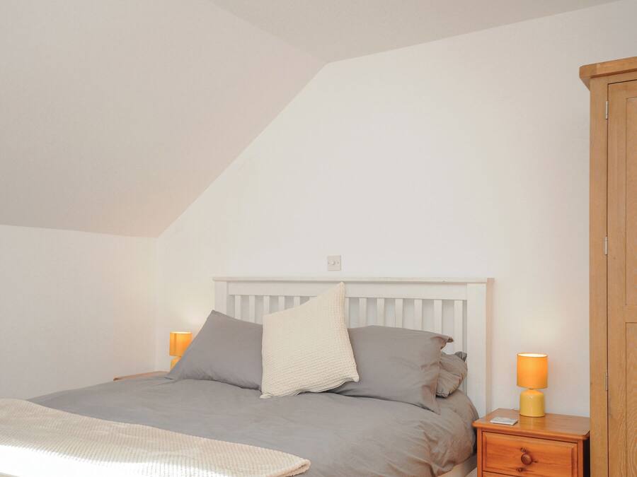 Een slaapkamer met een schuine balkenkap, een bed met grijze beddengoed, twee nachtlampjes en een houten garderobe.
