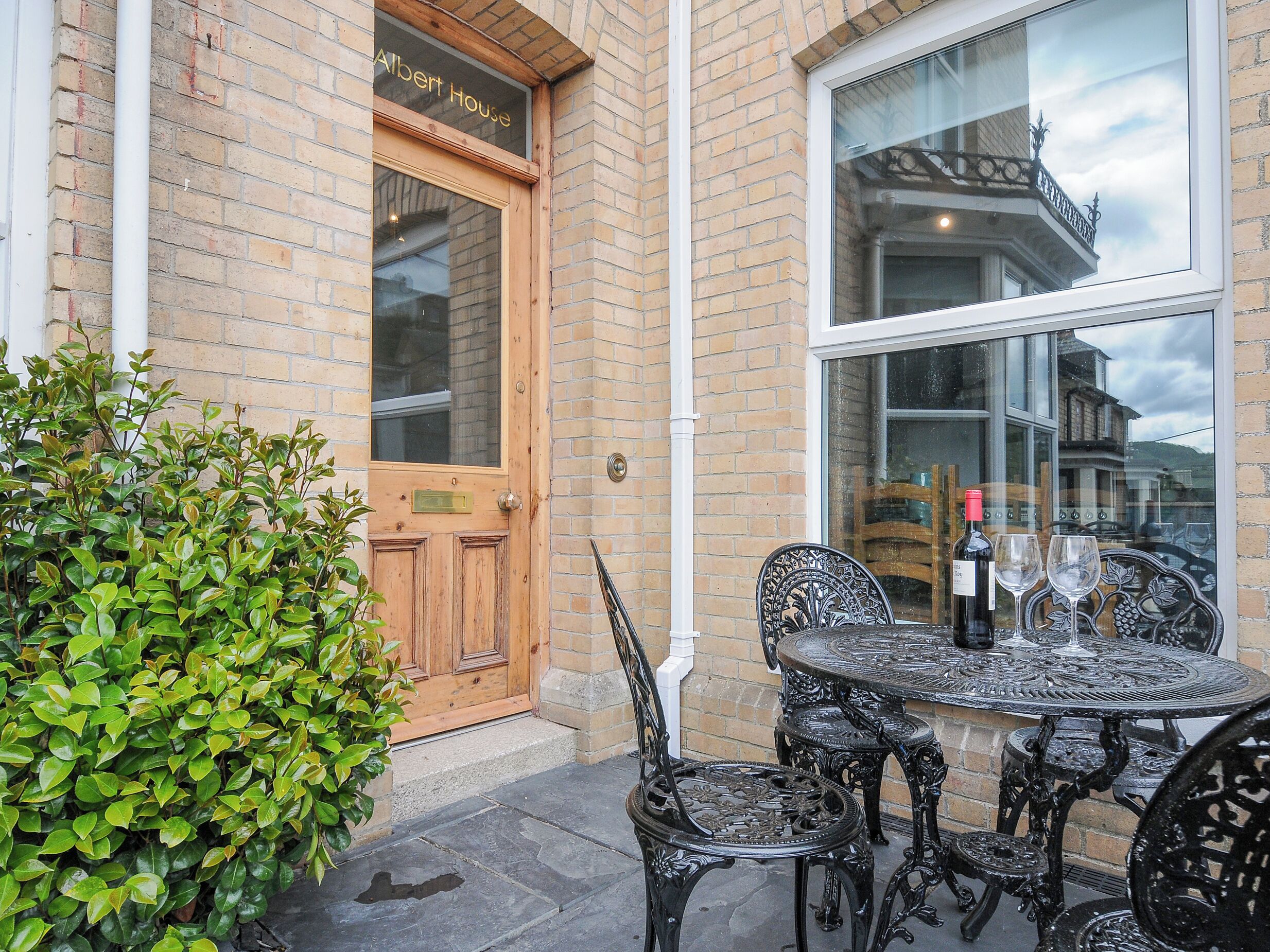 A brick building with a wooden door labeled "Albert House," a round metal table with chairs, and a window reflecting a building.