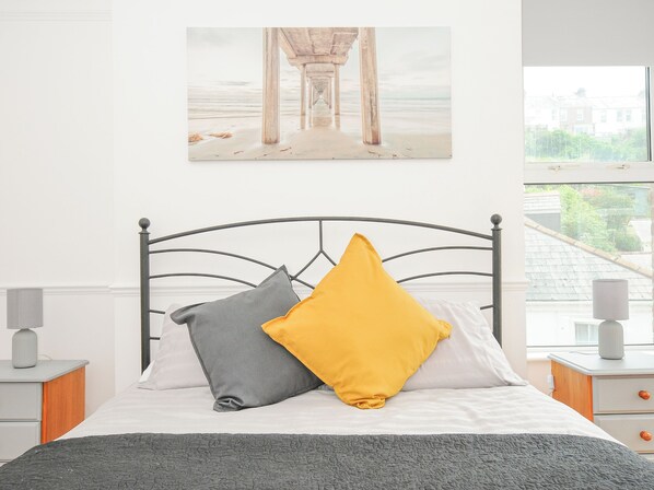 A neatly made bed with a grey and yellow pillow, a grey blanket, and a metal headboard. There is a painting of a pier on the wall above the bed. - Albert House (Looe)