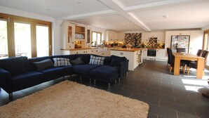 Een ruime zithoek met een donkerblauwe modulaire bank, een beige pluche vloerkleed en een keuken op de achtergrond met witte kasten en een betegelde spatwand.