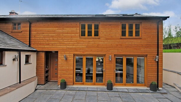 Een houten huis met twee verdiepingen, een stenen terras, grote glazen deuren en kleine ramen.