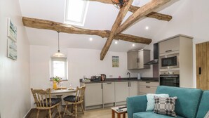 A cozy kitchen with wooden beams, a dining area with chairs, a teal sofa, and a small table with a remote control. - Y Stabl (Dyffryn Ardudwy)
