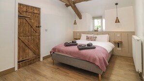 A bedroom with a sloped ceiling, wooden beams, a large bed with a pink blanket, two bedside tables, a wooden door, and a window. - Y Stabl (Dyffryn Ardudwy)
