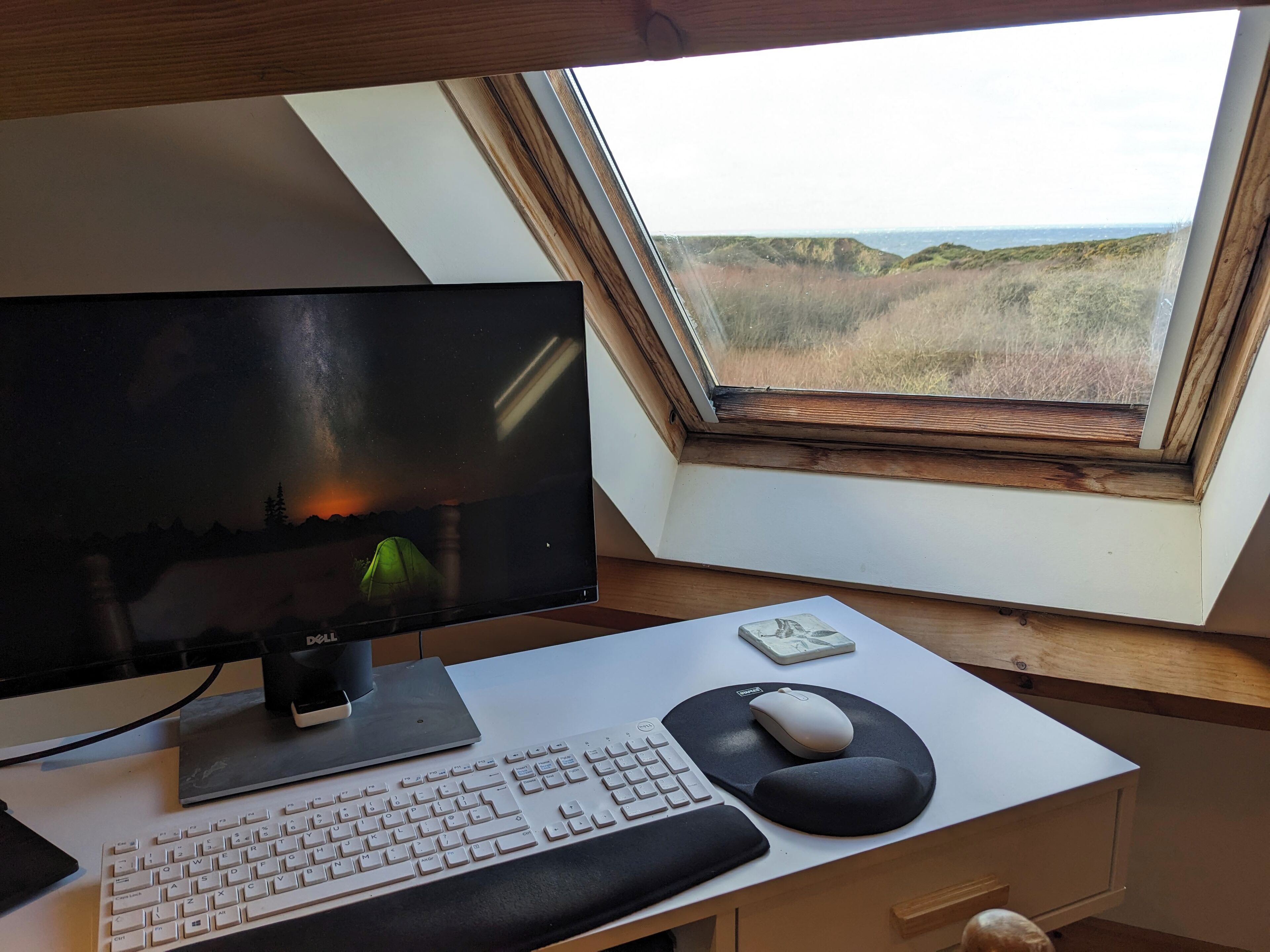 Un bureau d’ordinateur équipé d’un écran, d’un clavier, d’une souris, et offrant une vue sur un paysage grâce à une lucarne.