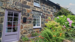 Une maison en pierre avec une porte et des fenêtres violettes, entourée d’un jardin où poussent différentes plantes et fleurs.