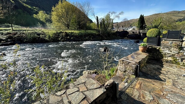 Un pont de pierre enjambe une rivière où l’eau coule, et on aperçoit, au loin, une colline et des arbres.
